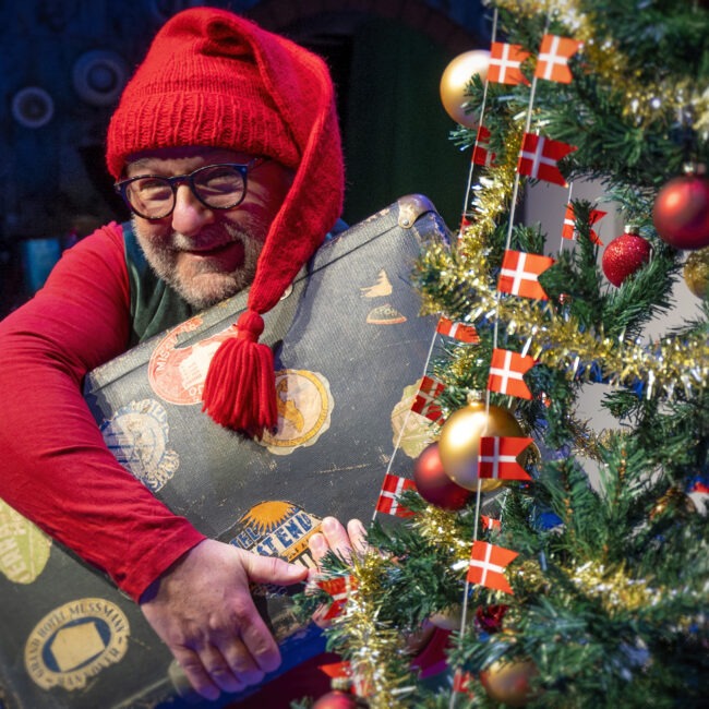 Billedet er en nisse med en kuffert bag et juletræ